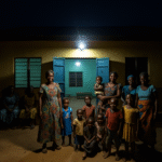 African women and children gathered at night under a solar-powered light outside a community centre.