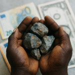 An African miner’s hands holding pieces of mineral ore, with blurred currency notes in the background under soft daylight.