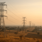 Aerial wide shot of transmission pylons crossing a dry African landscape at dawn, with a small village visible in the distance and warm golden light across the savannah.