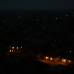 An aerial night-time photograph of an African town where only a few homes emit light, surrounded by widespread darkness, illustrating unreliable electricity access.