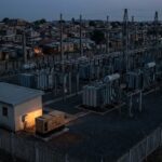 A dark electrical substation in an African city at night during a blackout, with dimly lit houses in the background and a single glowing generator in the foreground.